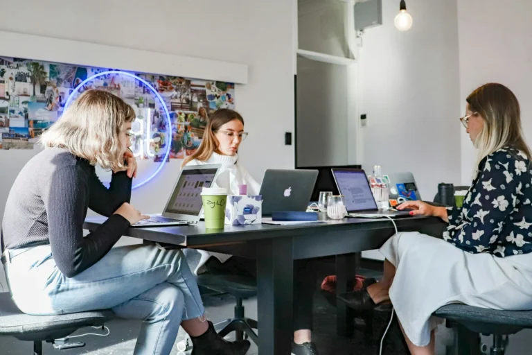 Three women working on laptops around a shared table in an office