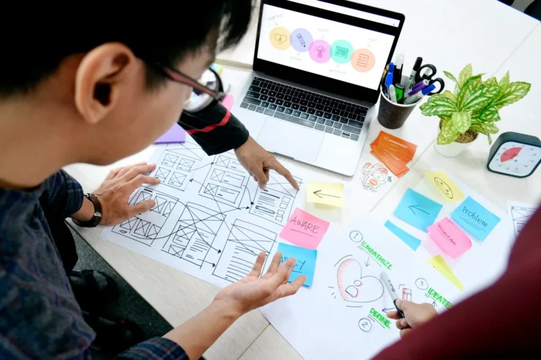 Designer sketching website wireframes at a desk beside a laptop during a website redesign