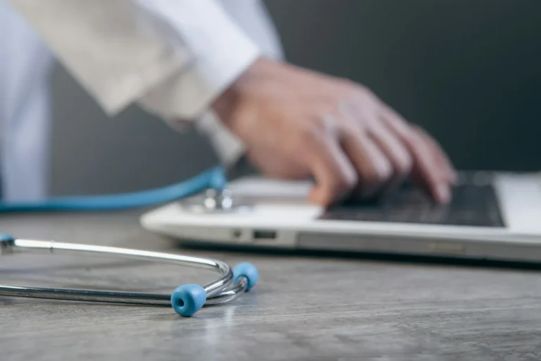 Doctor typing on a laptop beside a stethoscope while writing hospital website content
