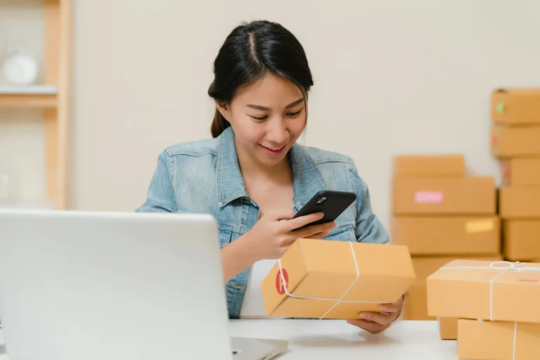 A smiling online shop owner checking a parcel on her phone beside a laptop